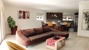 A modern living room with a brown sectional sofa, a yellow armchair, and a wooden coffee table. There is a kitchen in the background with a refrigerator, a sink, and a window with blinds. - Biko's Villa (Magaliesberg)