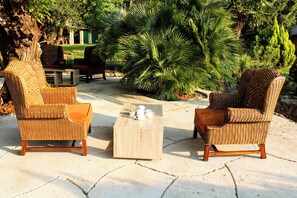 Deux fauteuils en osier avec des coussins, une table basse en bois, et en arrière-plan, des plantes tropicales et un bâtiment.