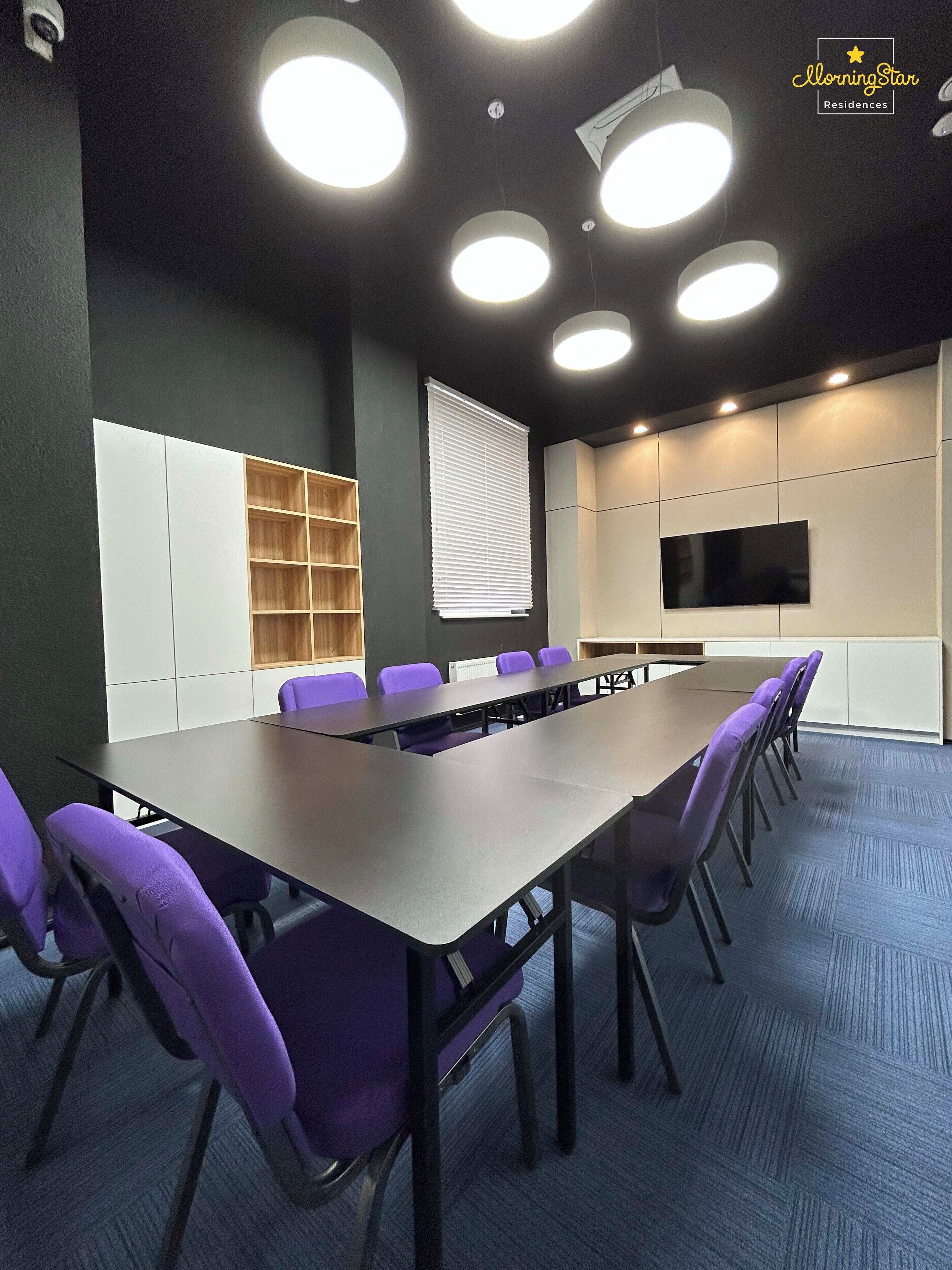Un salón de conferencias con una larga mesa, sillas moradas, un armario blanco, un televisor y luces en el techo.