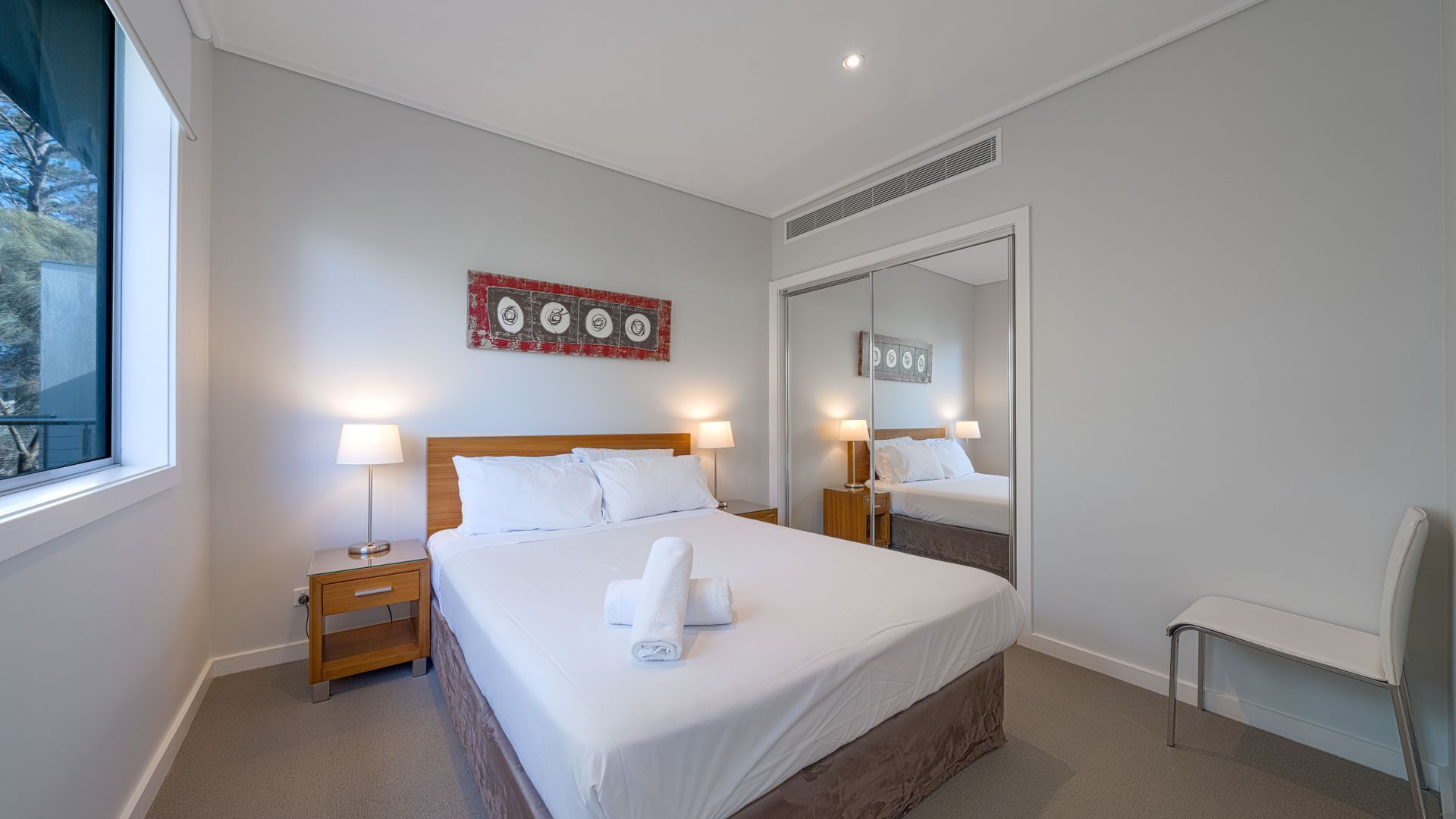 Une chambre d’hôtel avec un grand lit, deux tables de chevet avec des lampes, un miroir et une fenêtre donnant sur des arbres.