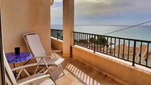 Un balcón con una silla blanca y una pequeña mesa redonda, con vistas a una playa y al mar.