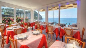 Uma área de refeições com mesas preparadas para hóspedes, grandes janelas com vista para o mar e uma varanda com guarda-corpo.