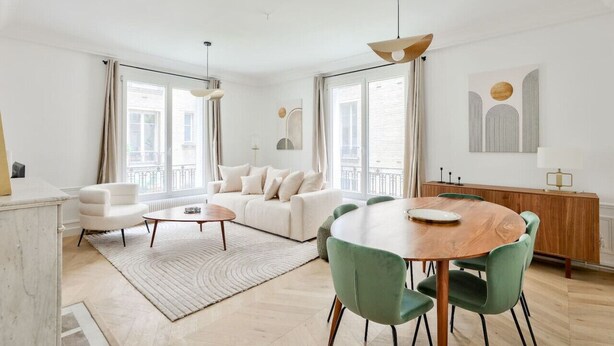 En moderne stue med en hvid sofa, et rundt spisebord med grønne stole, og store vinduer med gardiner.