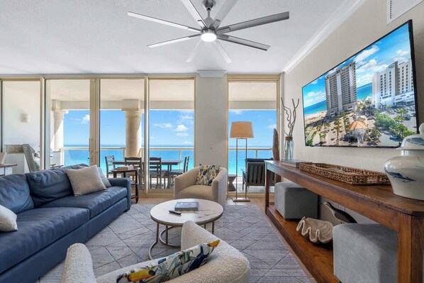 A spacious living room with a large TV, comfortable seating, a ceiling fan, and a balcony with a view of the ocean. - Beach Club 1105 - 𝑪𝒐𝒂𝒔𝒕𝒂𝒍 𝑹𝒆𝒕𝒓𝒆𝒂𝒕 (Pensacola Beach)