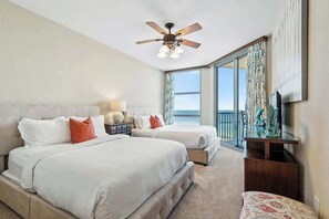 A hotel room with two beds, a ceiling fan, a nightstand with a lamp, a TV, and a view of the ocean.