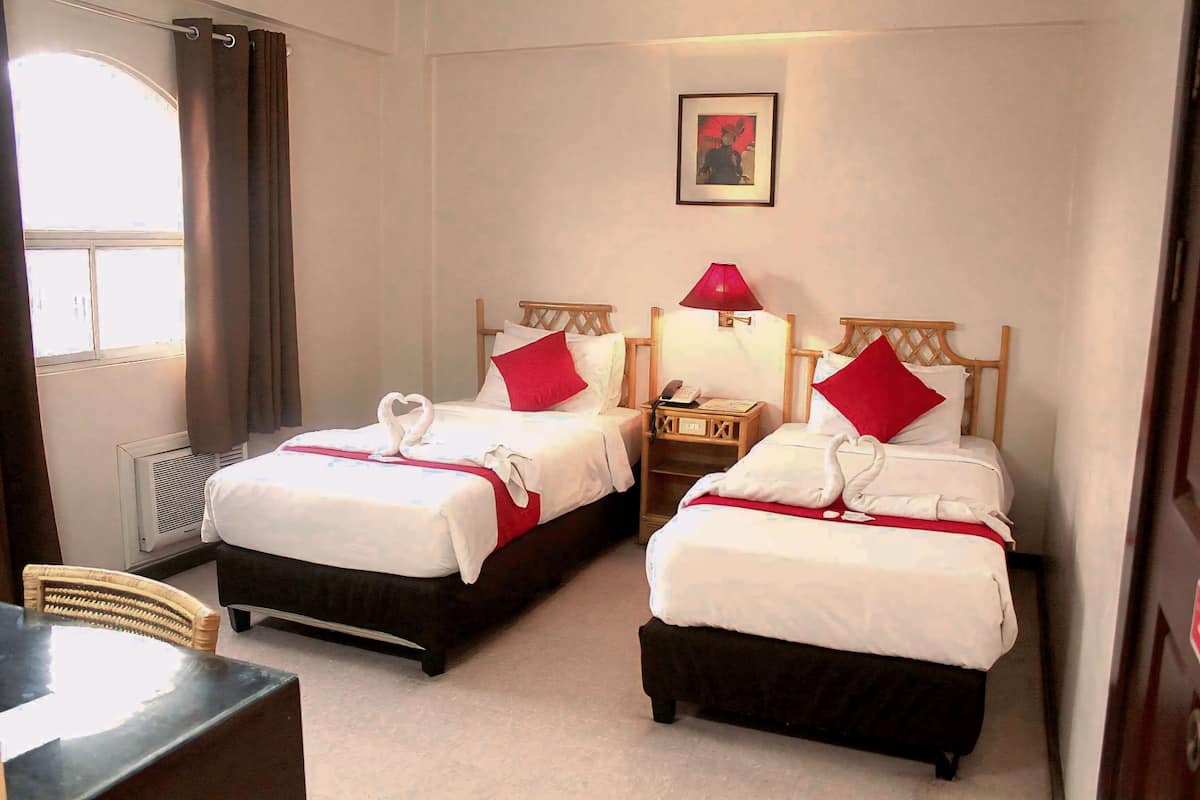 A hotel room with two beds, each with a red pillow and a white bedspread. There is a small table and a chair in the corner, and a window with curtains.