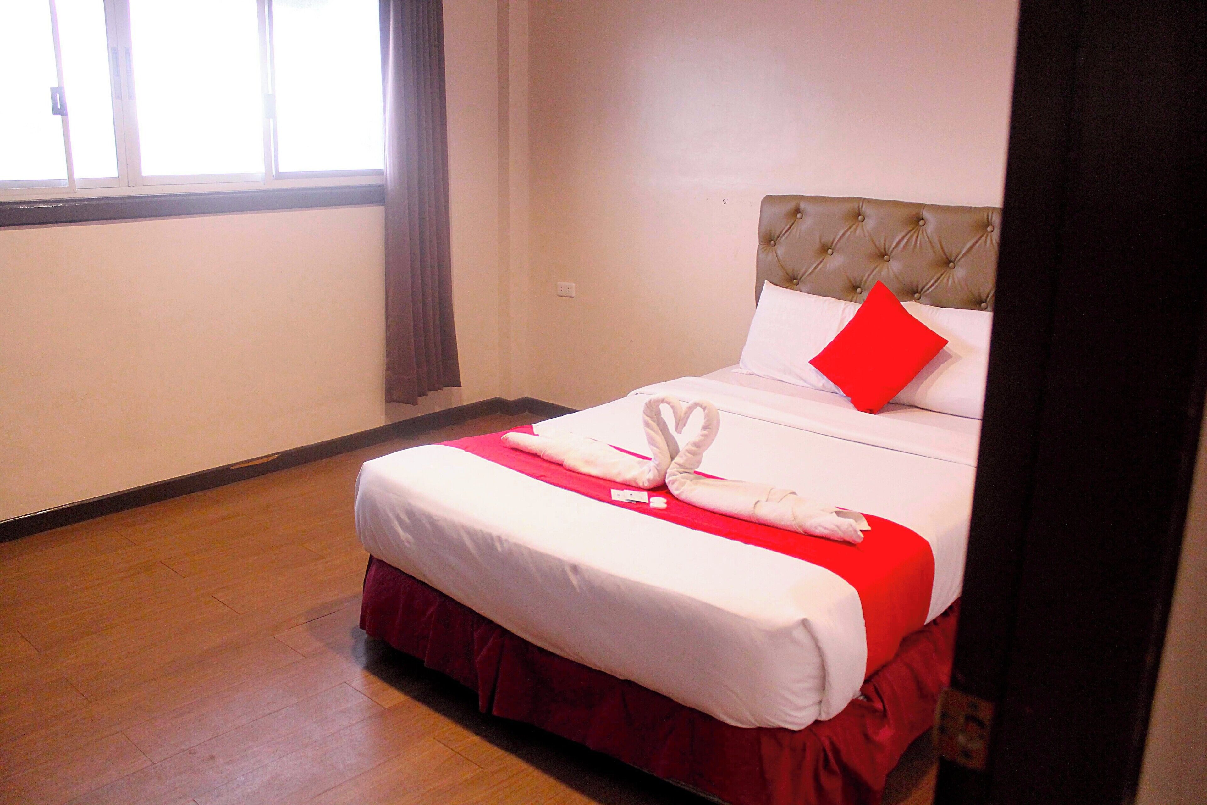 A neatly made bed with a red bedspread and a white pillow, a red accent pillow, and a decorative swan towel arrangement.