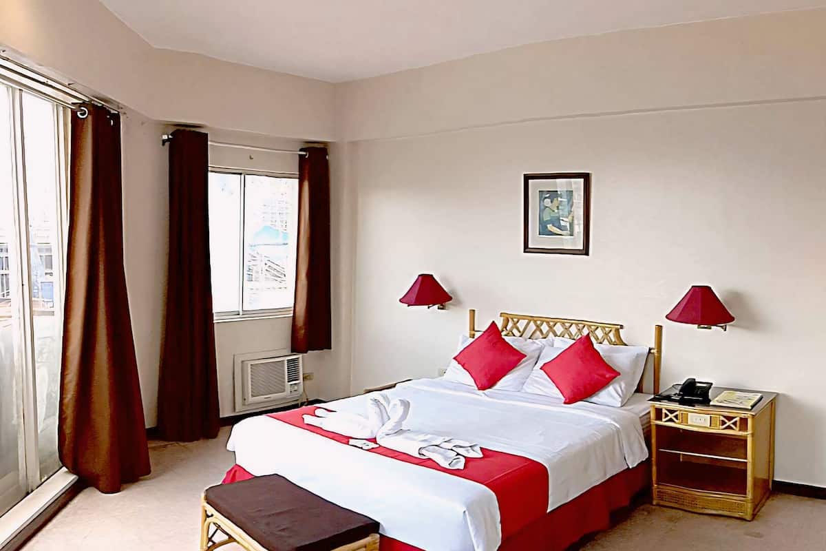 A hotel room with a large bed, a bedside table, a small stool, a window with curtains, and a wall-mounted lamp.