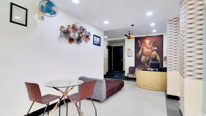 A reception area with a round glass table and two chairs, a sofa, a wall-mounted fan, a large painting, and a reception desk. - iROOMZ Growval Residency (Barasat)