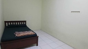 Een kleine eenpersoonskamer met een houten hoofdeinde, een donkergroen matras en een geborduurde plaid. De kamer heeft vloerbetegels en effen muren.