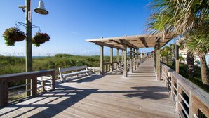 A wooden boardwalk with benches and hanging flower baskets, leading to a covered area with a view of the beach. - Adorable Beach Cottage,1 Block from Beach + Kure Beach Pier, Dog Friendly with Fence (Kure Beach)