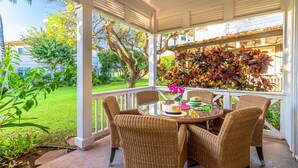 Un patio cubierto con sillas de mimbre y una mesa de vidrio, rodeado de exuberante vegetación y vistas a una zona residencial.