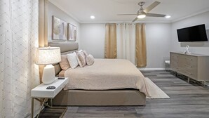 Een moderne slaapkamer met een bed, nachtkastje, lamp, gordijnen, plafondventilator, televisie en een dressoir.