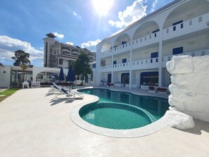 A swimming pool with a curved edge, a white building with multiple balconies, and a rock feature to the right.