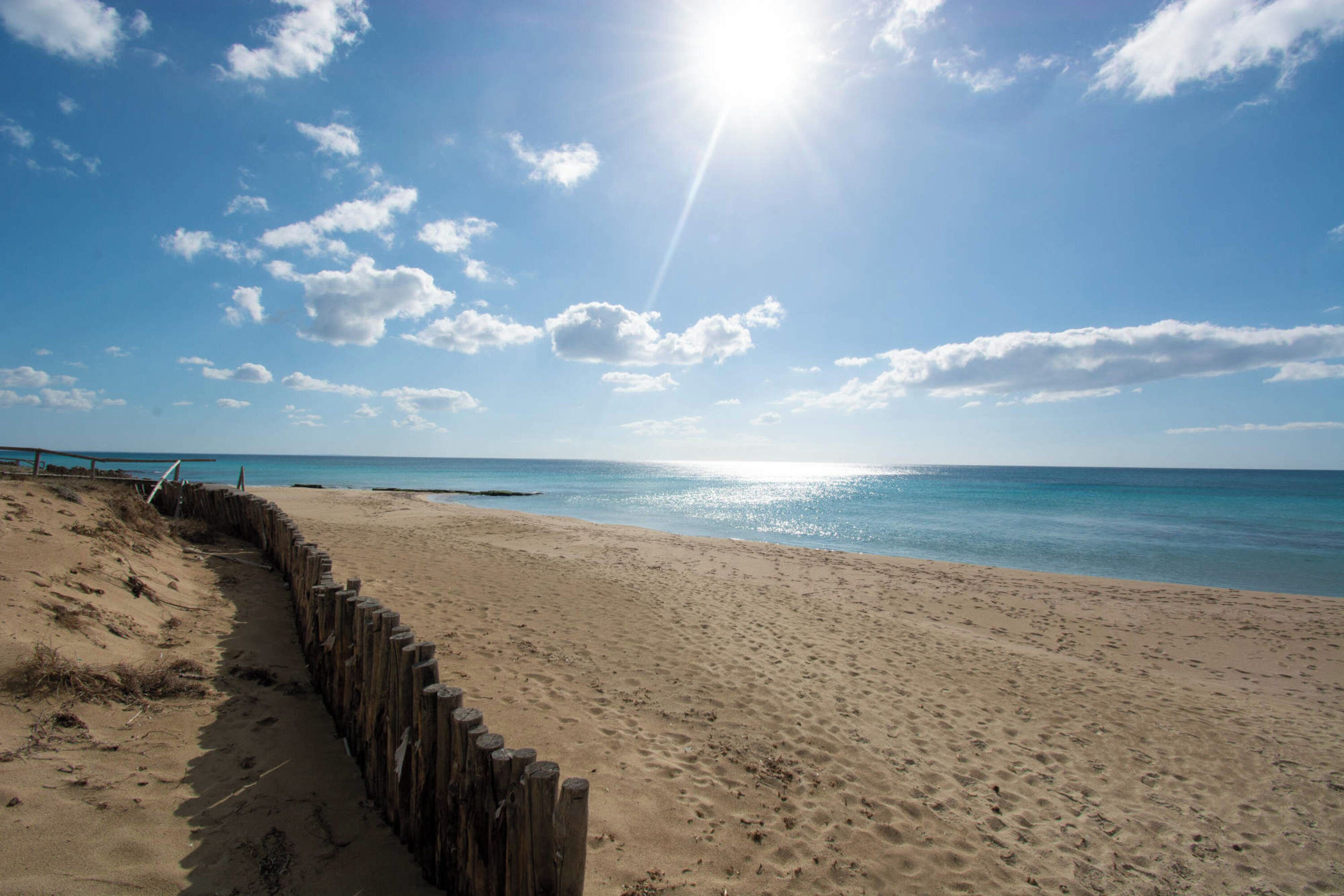 A sandy beach with a wooden fence, a clear blue sky, and the ocean in the background.