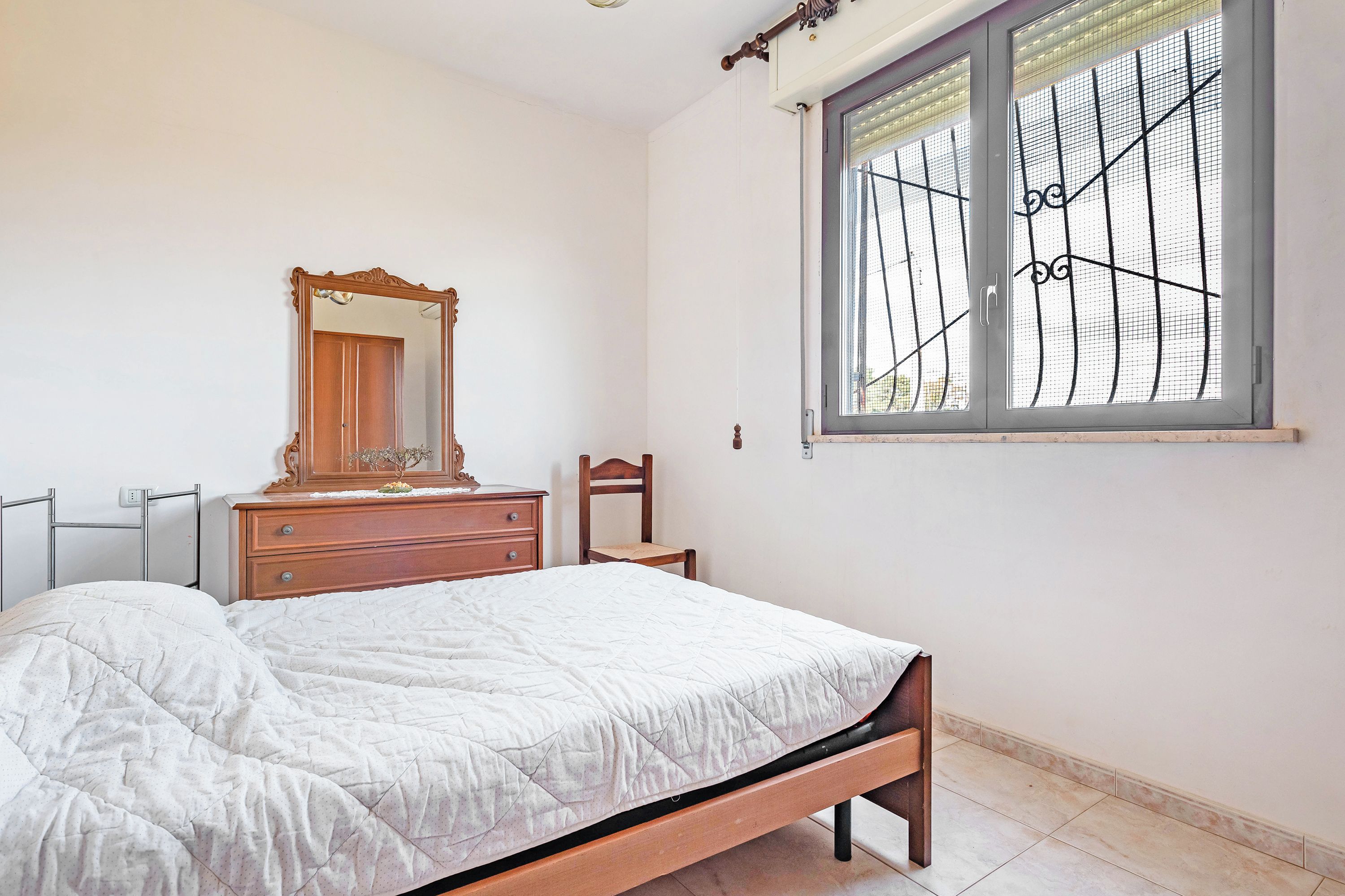 A bedroom with a bed, a dresser, a chair, a window with bars, and a mirror.