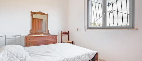 Quarto com cama, penteadeira, cadeira, janela com grades e espelho.
