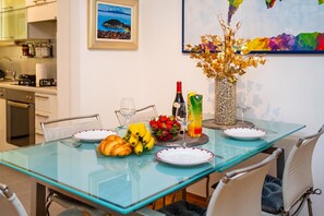 A dining table with a glass top, plates, a bottle, a vase with dried flowers, a bowl of fruit, and a croissant.