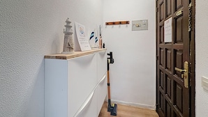 Un pasillo con pared blanca y texturizada, una puerta de madera, una estantería con un objeto decorativo, un certificado enmarcado y un enchufe en la pared.