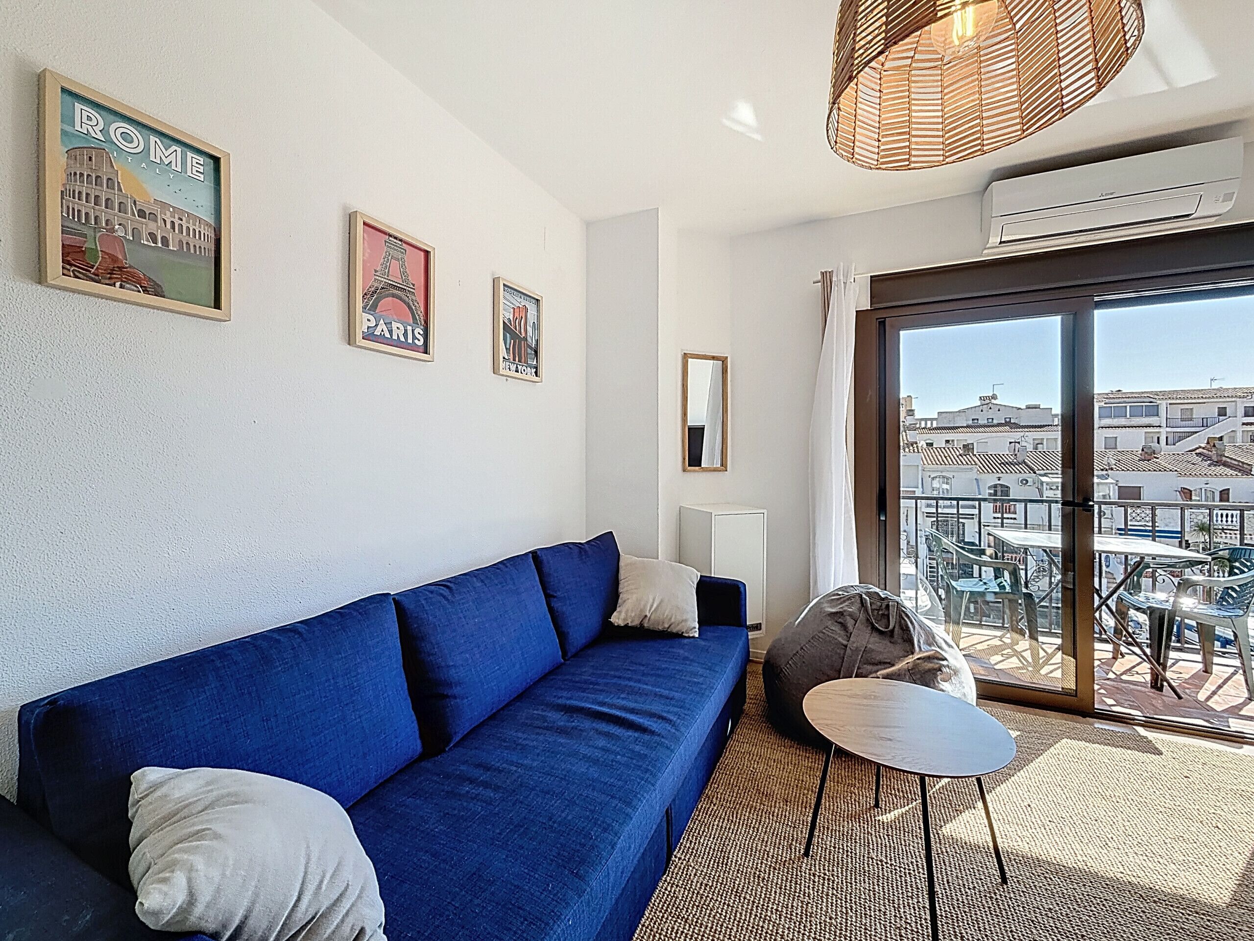 Una habitación con un sofá azul, una mesita redonda pequeña, una lámpara colgante y un ventanal con vistas a edificios.