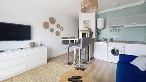 Una sala de estar moderna con un televisor de pantalla plana, un comedor con mesa y sillas, una cocina con lavadora y un sofá azul.