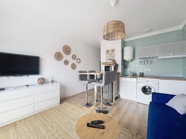 Un salon moderne doté d’un téléviseur à écran plat, d’un coin repas avec une table et des chaises, d’une cuisine équipée d’un lave-linge et d’un canapé bleu.
