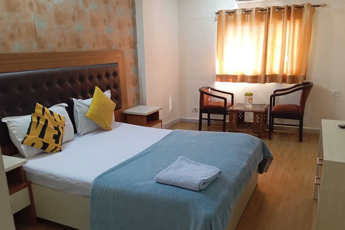 A neatly made bed with a blue blanket and pillows, a wooden headboard, a small table with a chair, and a window with curtains.