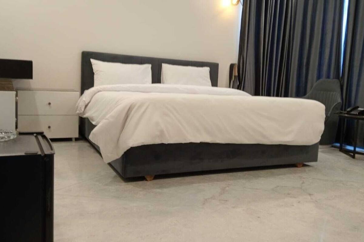 A modern hotel room with a large bed, a bedside table, a chair, and a small table with a lamp.