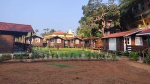 A row of wooden cabins with a central building, surrounded by a well-maintained garden and palm trees. - B R Cottages (Canacona)