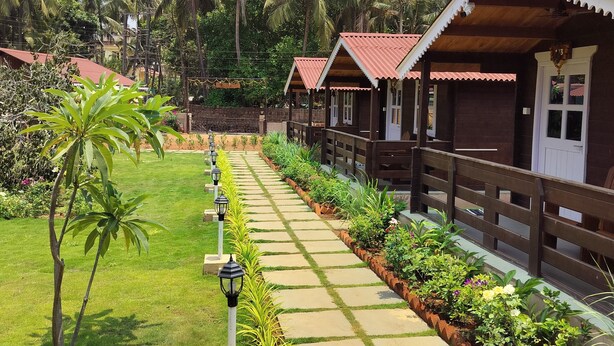 A pathway lined with small wooden cabins, surrounded by lush greenery and palm trees. - B R Cottages (Canacona)