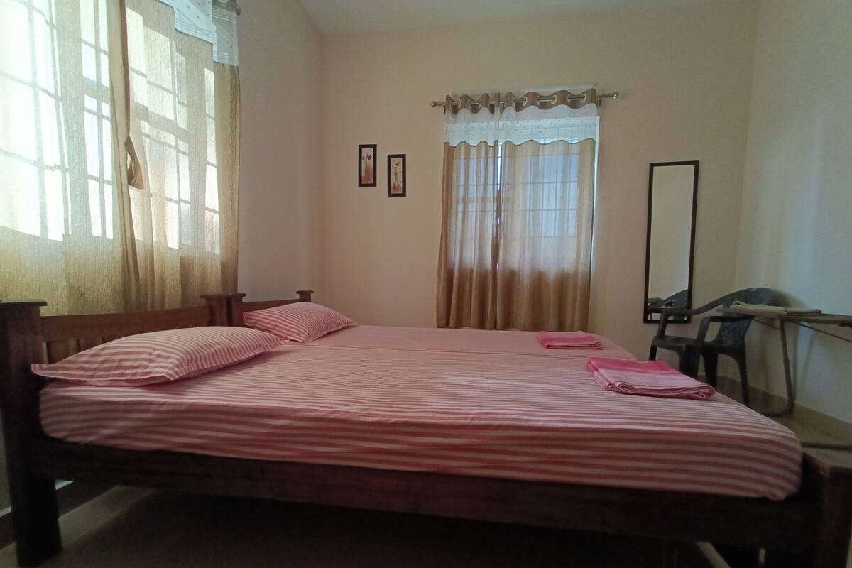 A single bed with a striped bedspread, a wooden headboard, and two pillows. There is a window with sheer curtains and a mirror on the wall.