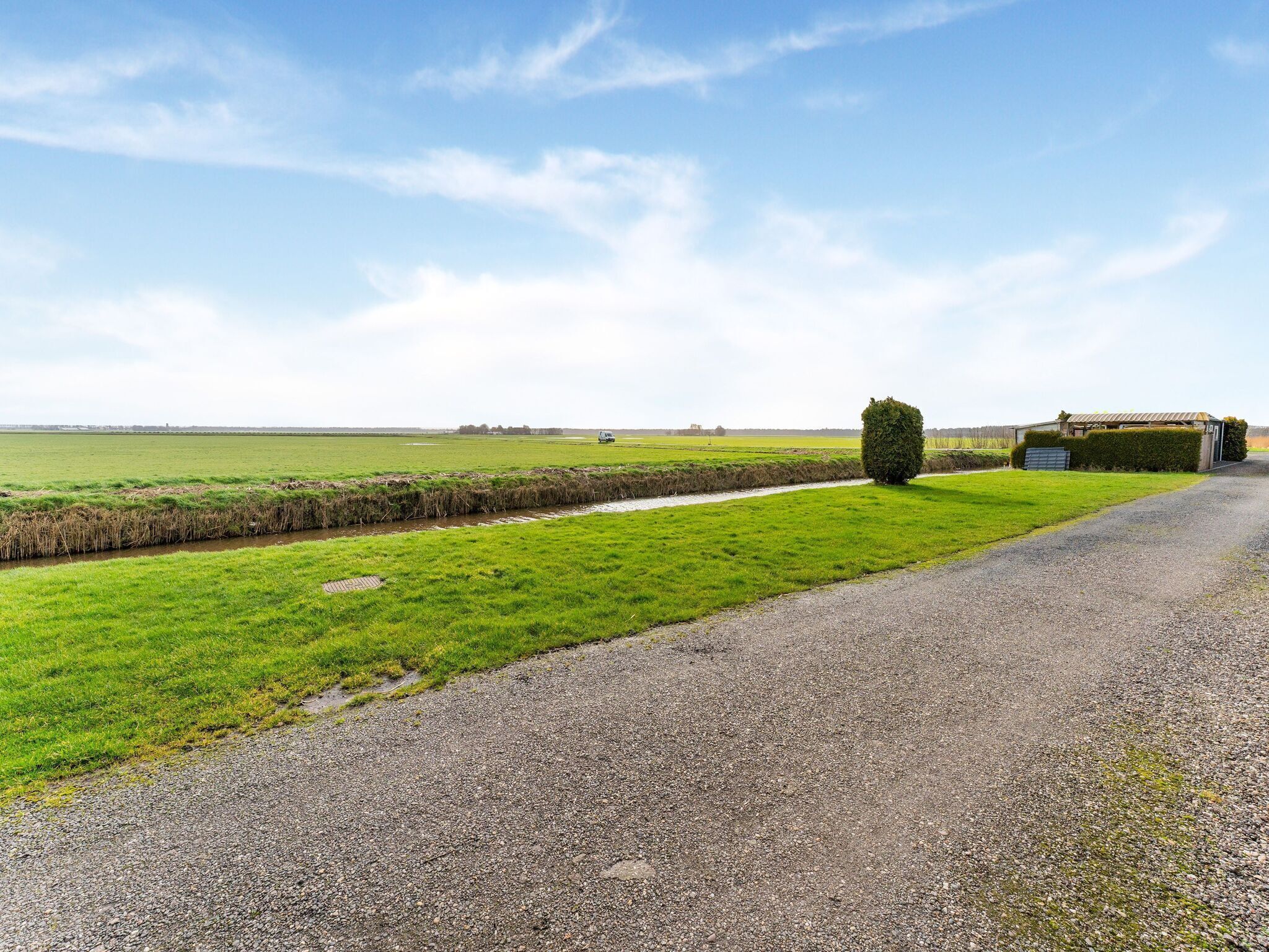 A rural road with a gravel path, a hedge, and a small building with a metal roof.