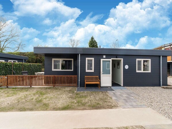 Une maison moderne à deux étages, avec une façade sombre, une terrasse en bois et un banc à l’extérieur.