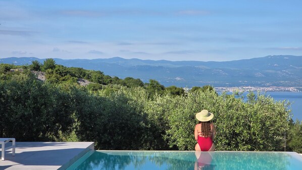 A woman in a red swimsuit and a straw hat sitting by an infinity pool with a view of a coastal city and mountains.