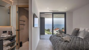Ein modernes Schlafzimmer mit einem großen Bett, einem Badezimmer mit Glasdusche und einem Balkon mit Meerblick.