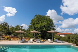A poolside area with lounge chairs and umbrellas, surrounded by trees and a clear sky.