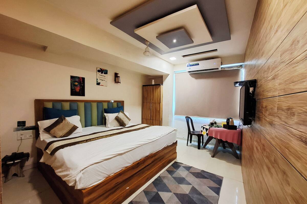Habitación de hotel con una cama de madera, un escritorio con mantel rojo, una silla y un televisor fijado a la pared.