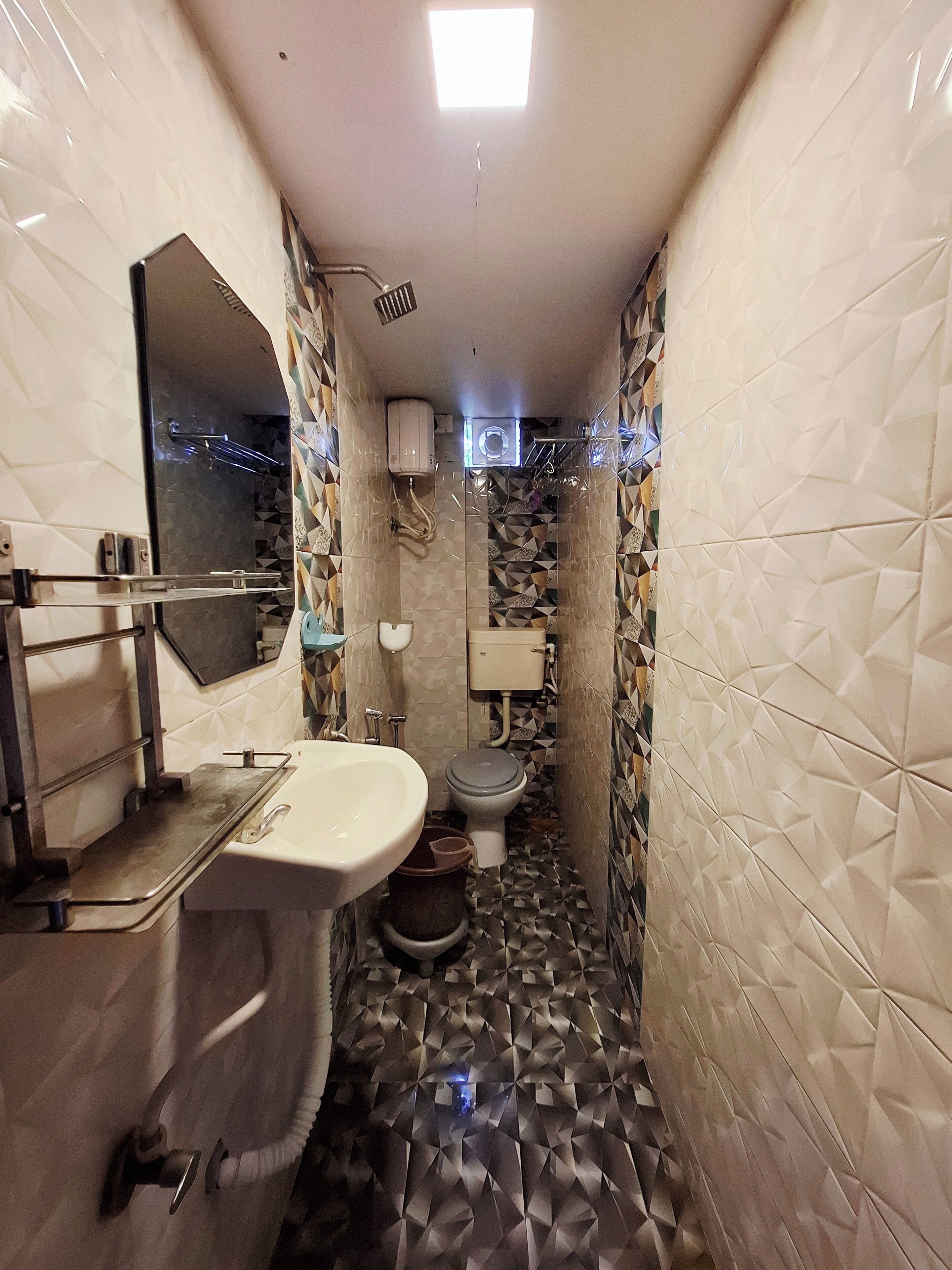 A narrow bathroom with a sink, toilet, and shower area. The walls are tiled with a geometric pattern, and there is a mirror above the sink.