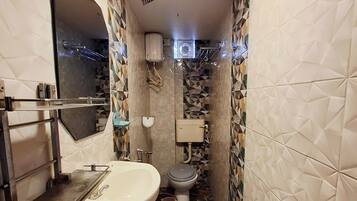 A narrow bathroom with a sink, toilet, and shower area. The walls are tiled with a geometric pattern, and there is a mirror above the sink.