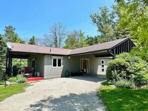 A single-story house with a covered porch, a gravel driveway, and a well-maintained lawn. - Porchside Retreat: Sauna and Treeline Backyard (Meaford)