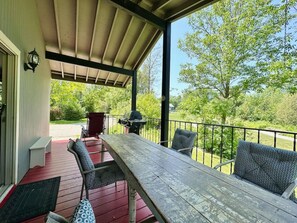 A covered outdoor patio with a wooden table and chairs, a barbecue grill, and a view of a green landscape. - Porchside Retreat: Sauna and Treeline Backyard (Meaford)
