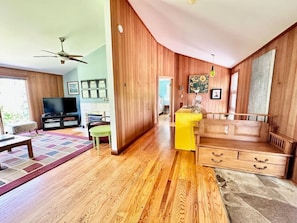 Uma sala de estar ampla com piso de madeira, ventilador de teto, televisão de tela plana, uma ilha de cozinha amarela e um banco de madeira.