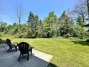 A backyard with two Adirondack chairs, a concrete patio, and a grassy lawn surrounded by trees. - Porchside Retreat: Sauna and Treeline Backyard (Meaford)