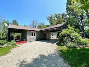 A two-story house with a covered porch, a gravel driveway, and a small garden area. - Porchside Retreat: Sauna and Treeline Backyard (Meaford)