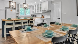 Cozinha moderna com armários brancos, mesa de jantar de madeira posta com tigelas e pratos azul-turquesa, e iluminação pendente.