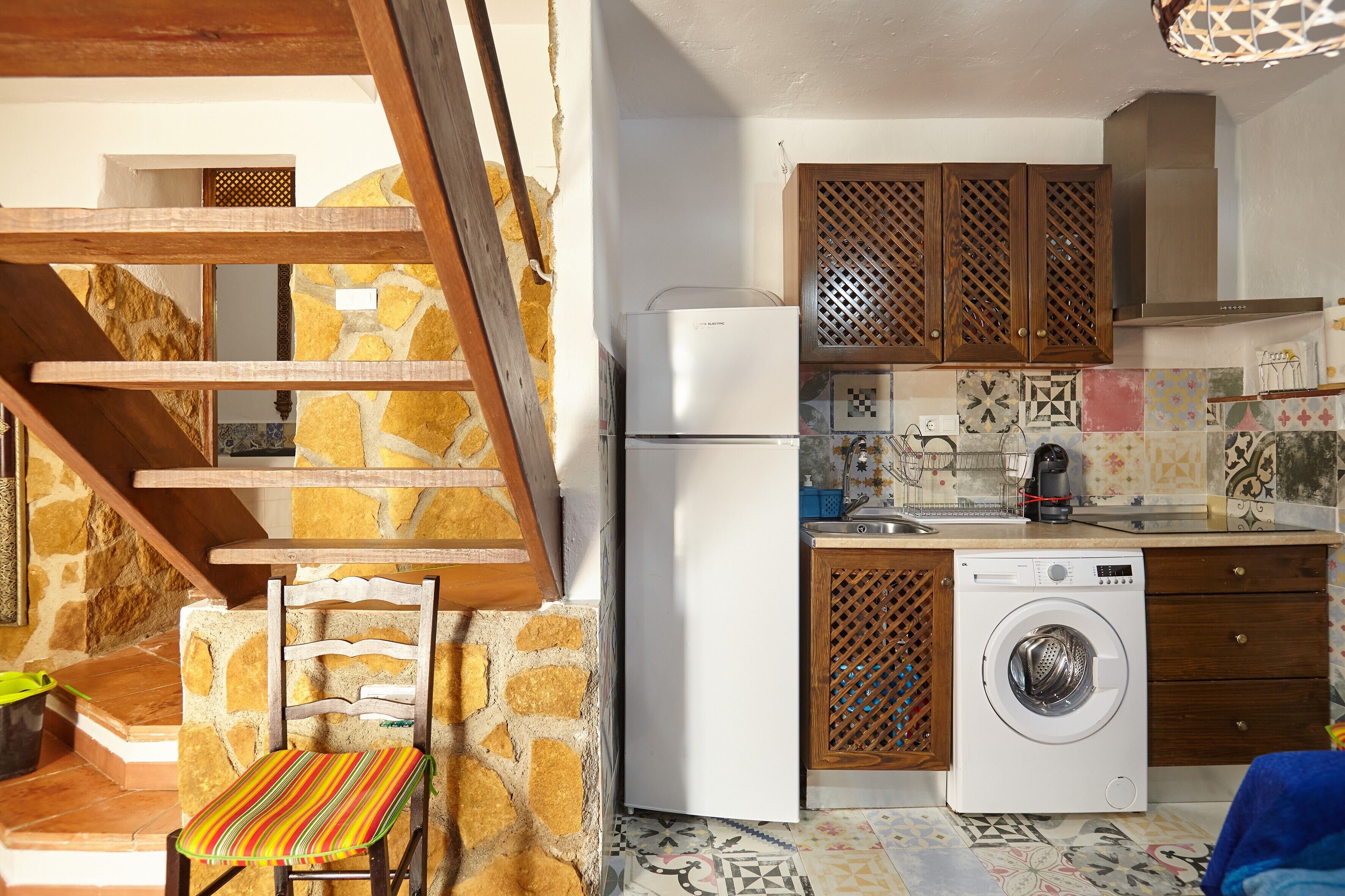 A kitchen with a washing machine, a refrigerator, wooden cabinets, and a chair.