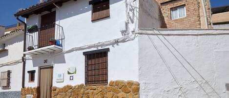 A white building with a stone base, a balcony with potted plants, a door with a window, and a small sign.