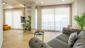 A modern living room with a grey sofa, a small round table, and a yellow armchair. There are large windows with sheer curtains, a flat-screen TV, and several potted plants.