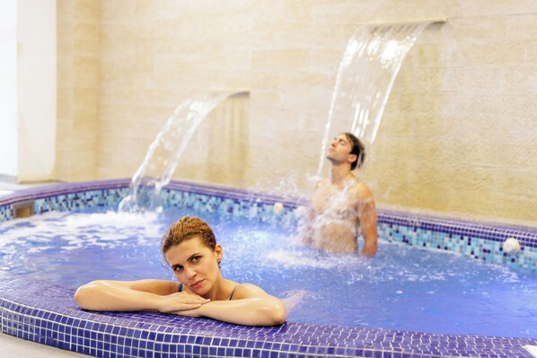 Un jacuzzi con dos personas, una sentada y otra de pie, con chorros de agua.