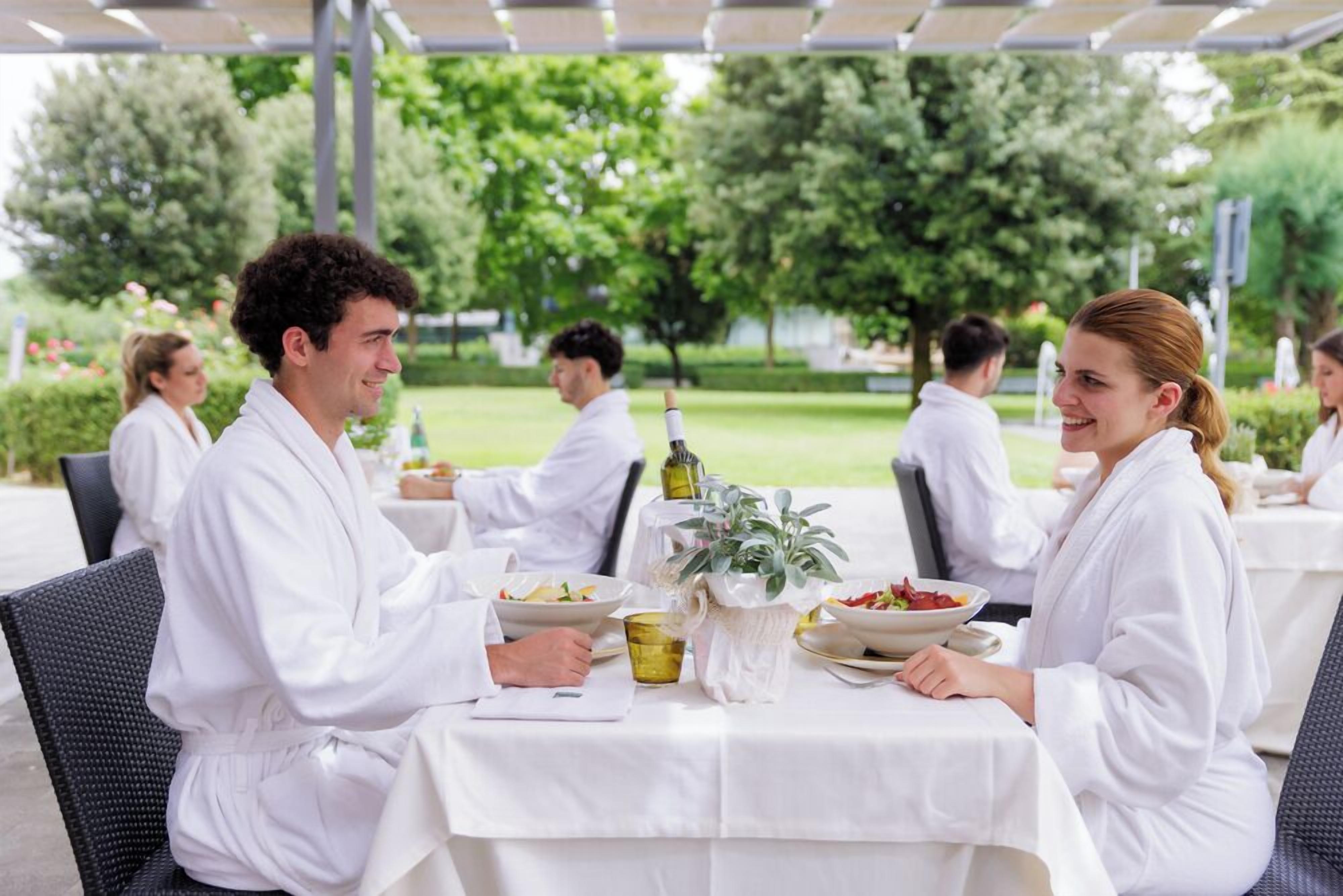 A group of people dining outdoors at a covered patio with white tablecloths and greenery in the background.